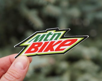 Op de afbeelding: Een sticker met de woorden "mtn BIKE" in groen en rood, omlijnd met zwart en geel. De sticker heeft een gekartelde, hoekige vorm en staat tegen een vage groene achtergrond.