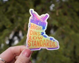 Op de afbeelding: Een holografische sticker in de vorm van een skischoen. De sticker heeft de tekst "High Dins Low Standards" in oranje en geel. Het schoenontwerp bevat paarse, roze en blauwe accenten.