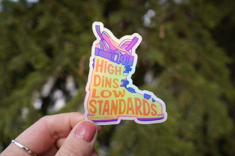 May include: A holographic ski boot sticker with the text "High Dins Low Standards". The boot design is in shades of purple, pink, and blue, with orange and yellow text. The sticker is a fun, eye-catching design.