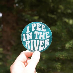 Può includere: Un adesivo rotondo con la scritta "I PEE IN THE RIVER" in bianco su sfondo turchese. Il testo è scritto in un carattere ondulato. L'adesivo è tenuto davanti a uno sfondo verde sfocato.
