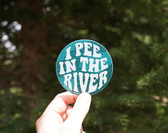 Op de afbeelding: Een ronde sticker met de tekst "I PEE IN THE RIVER" in witte, golvende letters op een turquoise achtergrond. De sticker wordt tegen een vage groene achtergrond van bomen gehouden.