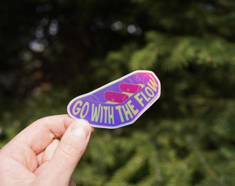 Op de afbeelding: Een sticker met een paarse achtergrond en de woorden "GO WITH THE FLOW" in een lichtgroen lettertype. De sticker heeft een bootachtige vorm met twee roze objecten erin. De sticker wordt in een hand gehouden.
