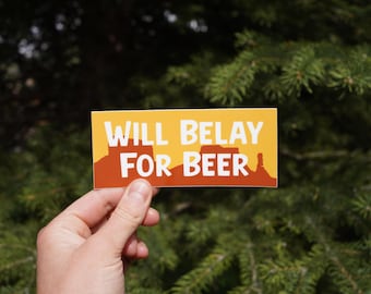 Op de afbeelding: Een rechthoekige sticker met de tekst "WILL BELAY FOR BEER" in het wit. De sticker heeft een gele achtergrond met een bruin en oranje landschapsontwerp. De sticker wordt door een hand vastgehouden.