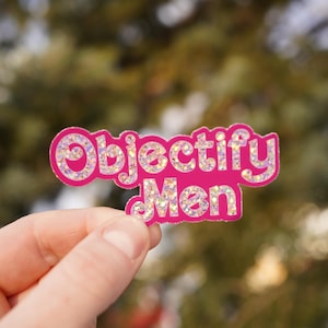 May include: Pink sticker with the text "Objectify Men" in white glitter letters.
