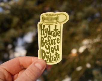 Op de afbeelding: Een gele sticker met een cartoontekening van een waterfles en de tekst "Hydrate Before You Dehydrate".