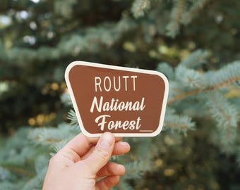 Op de afbeelding: Een bruine, trapeziumvormige sticker met een witte rand en de witte tekst ROUTT National Forest. Het ontwerp bootst een klassiek parkbord na met een schreefloos lettertype voor de bovenste regel en een cursief lettertype voor de rest.