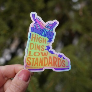 May include: A holographic sticker in the shape of a ski boot. The sticker has the words "HIGH DINS LOW STANDARDS" in a colorful, gradient font. The boot design features purple, blue, and yellow accents.