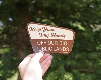 Op de afbeelding: Een bruin-witte sticker met de tekst "Keep Your Tiny Hands OFF OUR BIG PUBLIC LANDS". De sticker wordt vastgehouden voor een vage groene achtergrond van bomen.