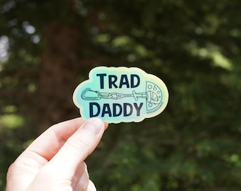 Op de afbeelding: Een holografische sticker met de woorden "TRAD DADDY" in zwarte blokletters. De sticker toont klimuitrusting, waaronder een karabijnhaak, een quickdraw en een cam. De achtergrond is een wazig groen.