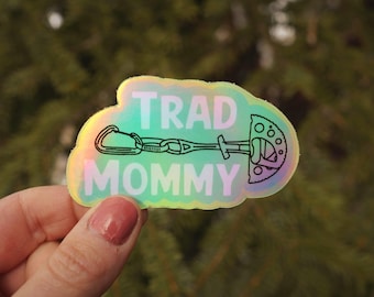 Op de afbeelding: Holografische sticker met de woorden "TRAD MOMMY" en een zwarte lijntekening van klimuitrusting. De sticker heeft een lichtgroen, blauw en roze iriserend effect. De sticker wordt voor een vage groene achtergrond gehouden.