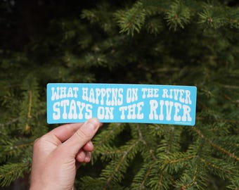 Op de afbeelding: Een lichtblauwe rechthoekige sticker met de witte tekst "WHAT HAPPENS ON THE RIVER STAYS ON THE RIVER", tegen een achtergrond van groene dennennaalden.
