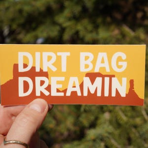 Puede incluir: Una pegatina amarilla y naranja con el texto "Dirt Bag Dreamin'" en blanco. La pegatina presenta una silueta de una cordillera.