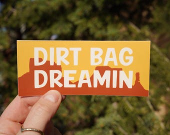 Op de afbeelding: Een gele en oranje sticker met de tekst "Dirt Bag Dreamin'" in wit. De sticker heeft een silhouet van een bergketen.