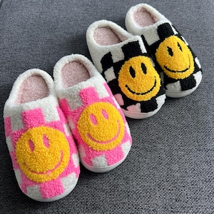 May include: Four pairs of plush slippers with smiley faces. Two pairs have a pink and white checkered pattern, and the other two have a black and white checkered pattern. Each slipper features a yellow smiley face.