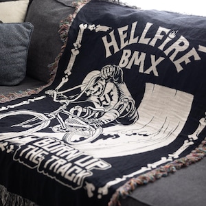 Puede incluir: Manta tejida en blanco y negro con un esqueleto montando una bicicleta BMX. La manta tiene el texto "Hellfire BMX" y "Burn Up The Track".