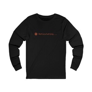 Puede incluir: Una camiseta negra de manga larga con un pequeño símbolo de asterisco naranja seguido del texto "Reticulating..." impreso en una fuente sans-serif sencilla en el centro del pecho. La prenda tiene un cuello redondo y mangas largas.
