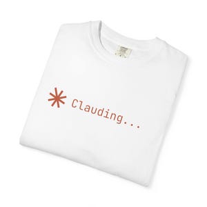 May include: A white short-sleeved t-shirt featuring a rust-coloured graphic on the chest. The graphic consists of an eight-pointed star symbol followed by the text "Clauding..." in a simple, sans-serif font. The shirt has a crew neckline.
