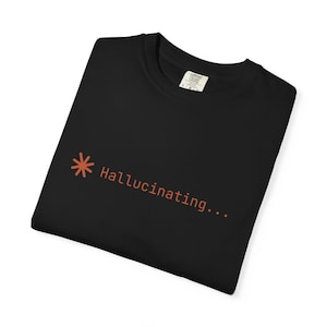 Könnte beinhalten: Ein schwarzes T-Shirt mit einem kleinen orangefarbenen Sternchen-Symbol, gefolgt von dem Text "Hallucinating..." in einer einfachen orangefarbenen Schriftart. Das Shirt hat einen Rundhalsausschnitt und ein sichtbares Innenetikett am Kragen.