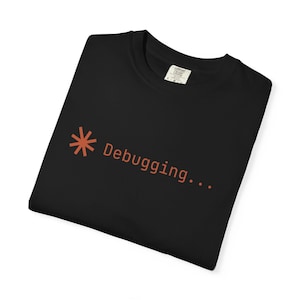 May include: Black t-shirt with the word "Debugging..." in orange. The design includes an asterisk symbol. The shirt is folded and appears to be made of a soft material.