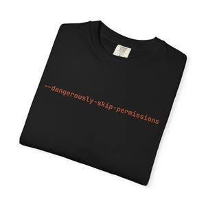 May include: A black t-shirt featuring the text --dangerously-skip-permissions printed in a rust-orange color across the chest. The shirt has a crew neckline and a visible interior neck label indicating a size large, presented on a flat surface.