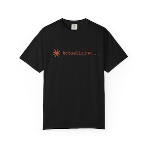 May include: Black t-shirt featuring the orange text "Actualizing..." with an asterisk symbol. The shirt is made of a soft material and has a crew neck. The design is simple and modern.