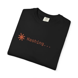 May include: Black t-shirt featuring the orange text "Hashing..." alongside an asterisk symbol. The t-shirt is folded, with a tag visible on the collar. The text and symbol are in a warm orange hue.