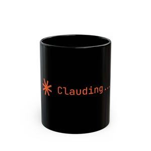 Könnte beinhalten: Ein schwarzer Keramikbecher mit einer orangefarbenen Grafik eines ladenden Sternsymbols, gefolgt von dem Text "Clauding..." in einer serifenlosen Schriftart. Der Becher hat eine zylindrische Form mit einer glatten, glänzenden Oberfläche.