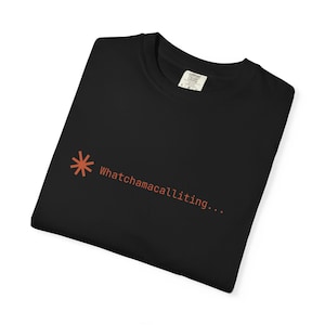 Puede incluir: Camiseta negra con el texto "Whatchamacallititing..." en naranja. La camiseta también presenta un símbolo de asterisco naranja. La camiseta está doblada y tiene una etiqueta en la esquina superior derecha.