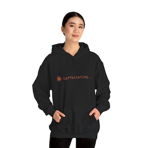 Op de afbeelding: Zwarte hoodie met een voorzak en trekkoord. De hoodie heeft de tekst "Caffeinating..." in oranje, samen met een oranje asterisk-symbool. De hoodie heeft een casual stijl.