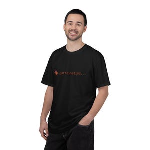 May include: Black t-shirt with the text "Caffeinating..." in orange. The text is above a stylized orange sunburst graphic. The shirt is worn by a person.