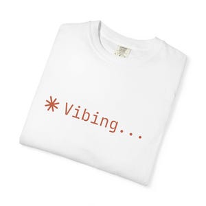 Puede incluir: Una camiseta blanca de cuello redondo con un símbolo de asterisco de color óxido seguido del texto "Vibing..." impreso en una fuente sans-serif sencilla en el pecho. La prenda incluye una etiqueta interior visible con detalles de la marca.
