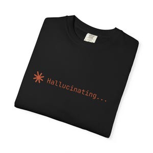 Puede incluir: Una camiseta negra de cuello redondo con un pequeño símbolo de asterisco naranja seguido del texto "Hallucinating..." impreso en una fuente sans-serif sencilla en el lado izquierdo del pecho. La prenda tiene una etiqueta interior visible.