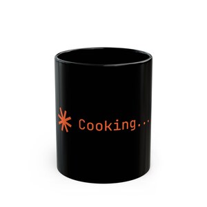 Könnte beinhalten: Ein schwarzer Keramikbecher mit einer orangefarbenen Grafik eines ladenden Sternchensymbols, gefolgt von dem Text "Cooking...", der in einer klaren serifenlosen Schrift auf der Seite des zylindrischen Gefäßes mit glatter Oberfläche gedruckt ist.