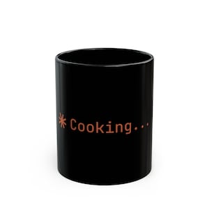 May include: Black ceramic mug with the word "Cooking..." in orange text. The mug has a simple, cylindrical shape and a glossy finish. The text is in a sans-serif font.