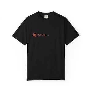 May include: Black t-shirt with short sleeves. The shirt features a small orange asterisk symbol and the word "Musing..." printed in orange. The shirt is a solid color and has a simple, minimalist design.