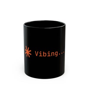 May include: A black ceramic mug featuring an orange graphic of a starburst icon followed by the text "Vibing..." printed in a simple sans-serif font on the side. The mug has a smooth, glossy finish and a cylindrical shape with a flat base.