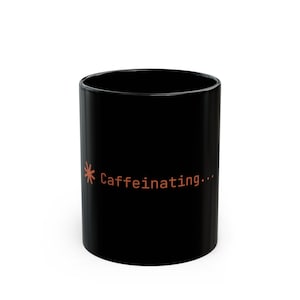 May include: Black coffee mug with the word "Caffeinating..." in orange text. The mug has a simple, cylindrical shape and a glossy finish. The text is accompanied by a small asterisk-like design.