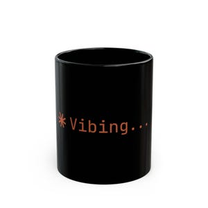 May include: A black ceramic mug with the text "Vibing..." in burnt orange. The mug is cylindrical and has a glossy finish. The text is in a sans-serif font. A simple, everyday item.