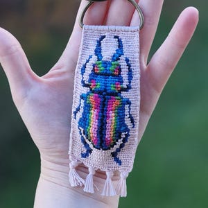 Pode incluir: Chaveiro artesanal com um desenho de besouro bordado em cores do arco-íris. O besouro está sobre um fundo rosa claro, com pequenas borlas na parte inferior. Um anel de metal é preso para as chaves.