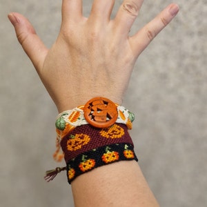 Op de afbeelding: Een verzameling handgemaakte vriendschapsarmbanden met een Halloween-thema. De armbanden hebben oranje pompoenpatronen, een houten pompoenknop en diverse kleuren, waaronder oranje, wit, groen en bordeauxrood.