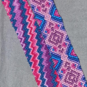 Może przedstawiać: Dwa bransoletki przyjaźni z tkanymi nitkami w kolorze różowym, fioletowym i niebieskim. Bransoletki mają różne wzory i są ze sobą związane na końcach.