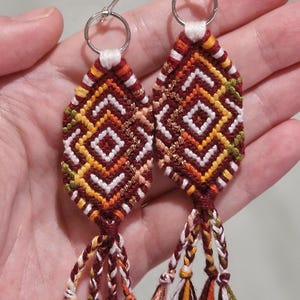 Op de afbeelding: Een paar handgemaakte, geweven oorbellen met een ruitpatroon in tinten bordeauxrood, geel, wit en groen. De oorbellen hebben zilverkleurige ringen en gevlochten kwastjes in bijpassende kleuren. De oorbellen zijn in bohemian stijl.