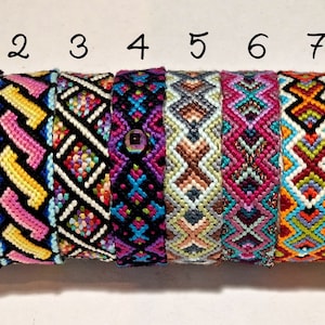 以下が含まれることがあります： 異なるパターンを持つ8本のカラフルな友情ブレスレットが、白い糸の巻き取り機に巻き付けられています。ブレスレットは1から8まで番号が付けられています。