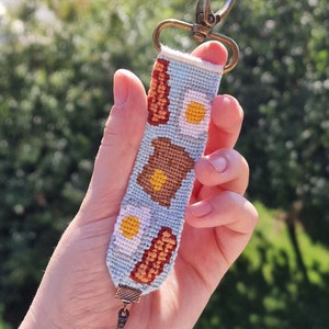 Può includere: Un portachiavi intrecciato blu e marrone a tema colazione. Il portachiavi presenta un uovo fritto, pancetta, toast e un piccolo ciondolo a forma di padella.