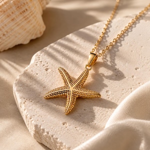 May include: A gold-toned necklace featuring a textured starfish pendant with a detailed, bumpy surface. The pendant is attached to a delicate gold-coloured chain, showcasing a realistic five-pointed sea creature design with a polished metallic finish.