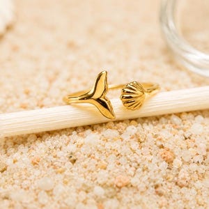 May include: A gold-toned open ring featuring a whale tail charm on one end and a small seashell charm on the other. The metal band is polished and reflective, showcasing a smooth finish on the tail and ribbed detailing on the seashell design.