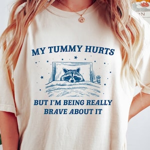 May include: A cream coloured t-shirt with a blue graphic of a raccoon in bed with the text "My tummy hurts but I'm being really brave about it".
