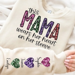 Peut inclure: Un sweat-shirt crème avec le texte "this MAMA wears her heart on her sleeve" en lettres arc-en-ciel scintillantes. Les manches ont trois cœurs scintillants avec les noms "Emilya", "Jamie" et "Sophie" écrits en dessous.