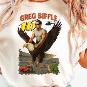 May include: Cream t-shirt with a graphic of a man riding an eagle over a race track. The text "GREG BIFFLE 16" is above the image. A helicopter is also depicted in the background.