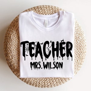 May include: A white t-shirt featuring the word "TEACHER" in a black, dripping font, with Halloween-themed decorations. The name "MRS. WILSON" is printed underneath. The shirt is folded and placed on a woven, circular surface.